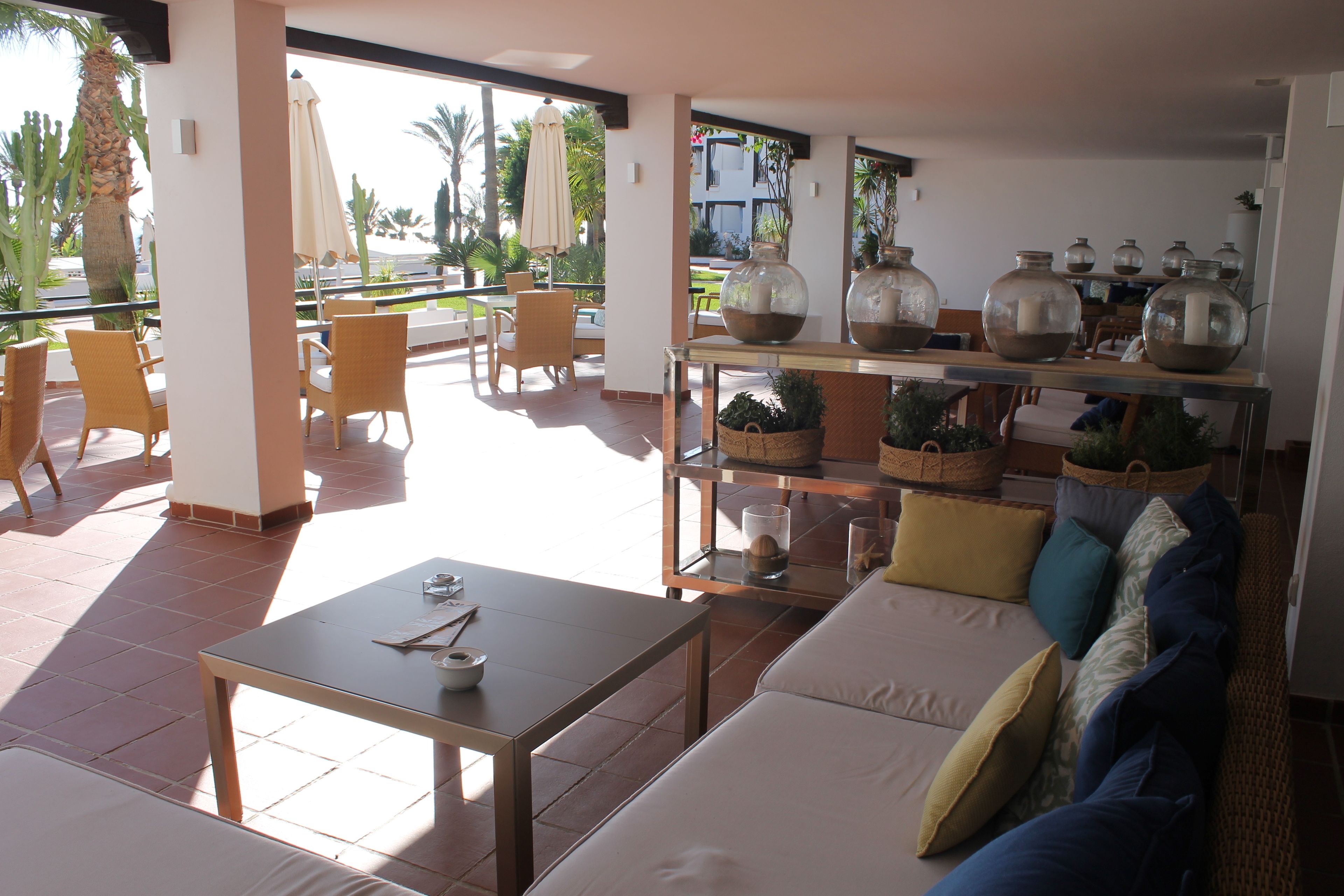 A spacious living area with a sofa, a coffee table, and a dining table. There are potted plants and vases on the table. The room has large windows with a view of palm trees and a building.