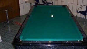 A pool table with a green felt surface, a white ball, and surrounding chairs.