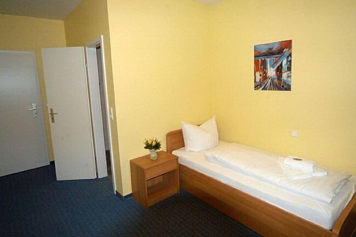 Una habitación pequeña con una cama individual, cabecera de madera, mesita de noche con un florero y un cuadro en la pared.