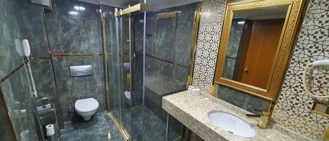 Un baño moderno con ducha envuelta en vidrio, inodoro, lavamanos con espejo de marco dorado y pared con diseño.