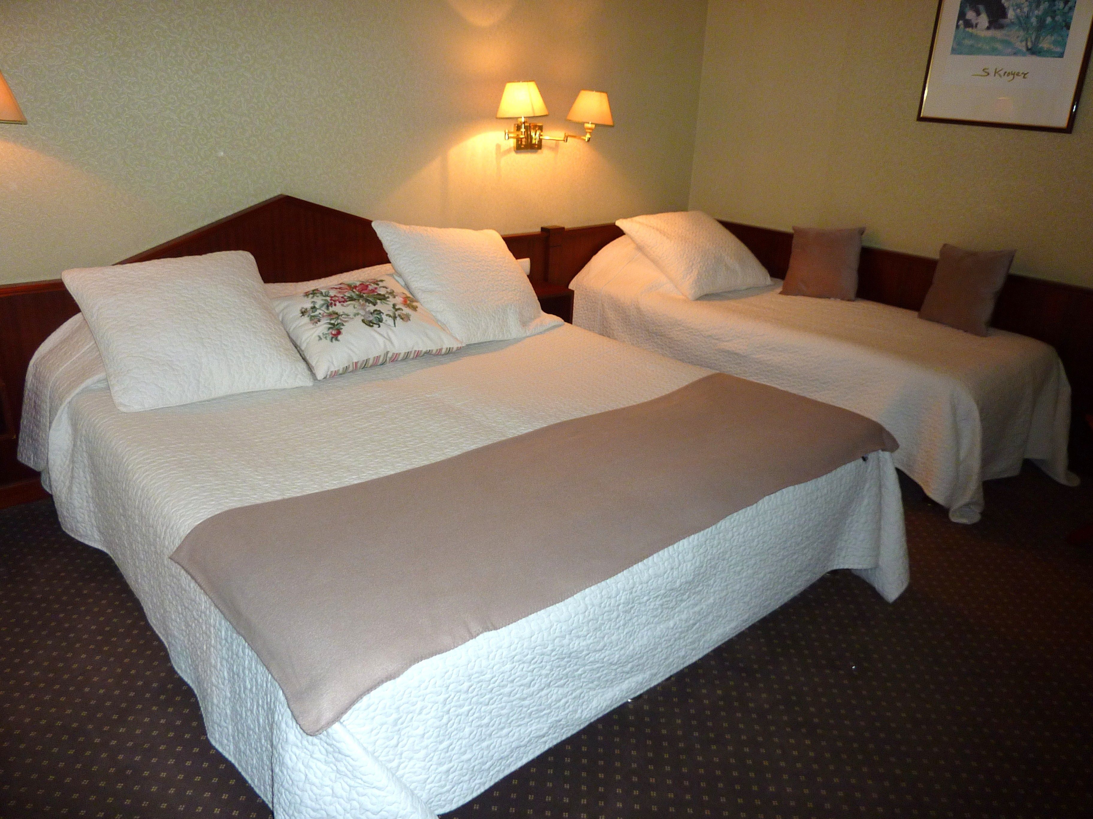 A hotel room with two beds, each with white bedding and a brown throw blanket. There are two wall-mounted lamps providing light.