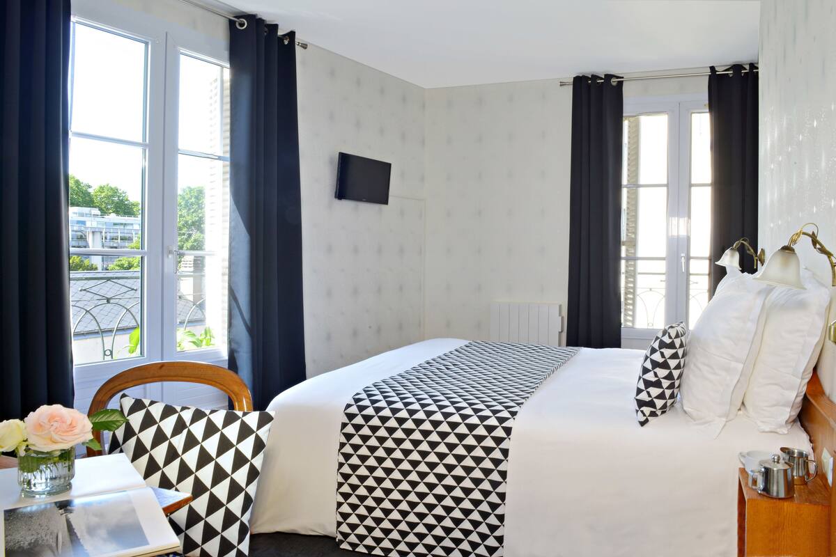 A hotel room with a bed, a chair, a table with a vase of flowers, and a window with curtains.
