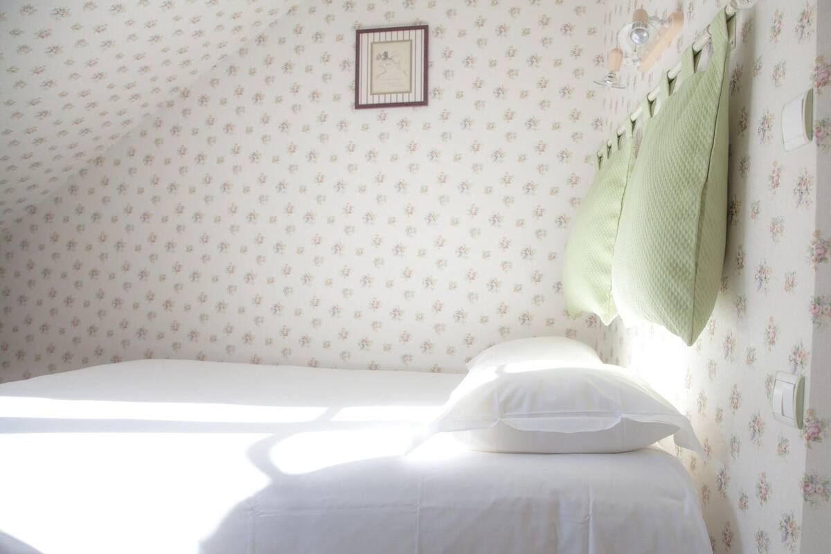 A bedroom with a single bed, floral wallpaper, a framed picture on the wall, and a green towel hanging on the bed.