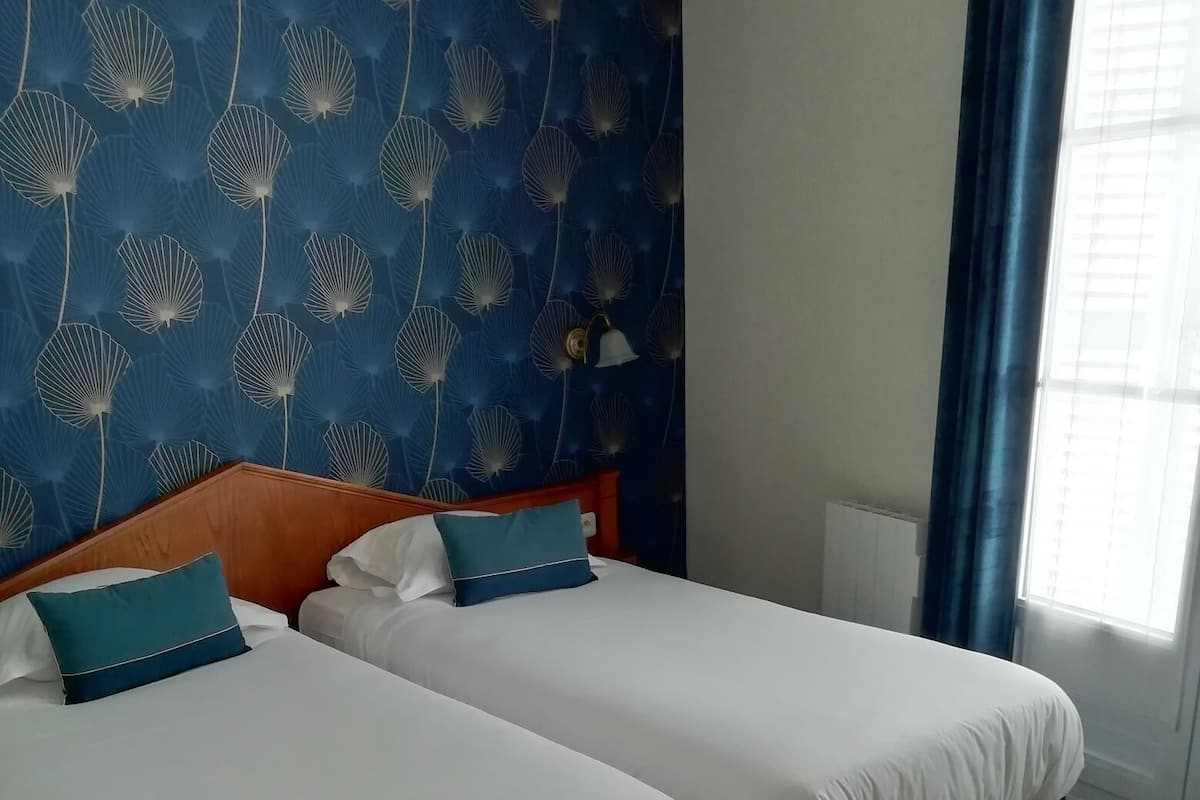 A hotel room with two beds, a blue patterned wall, and a window with curtains.