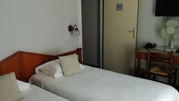 A hotel room with two single beds, a wooden headboard, a desk with a chair, a television, and a wall-mounted fan.