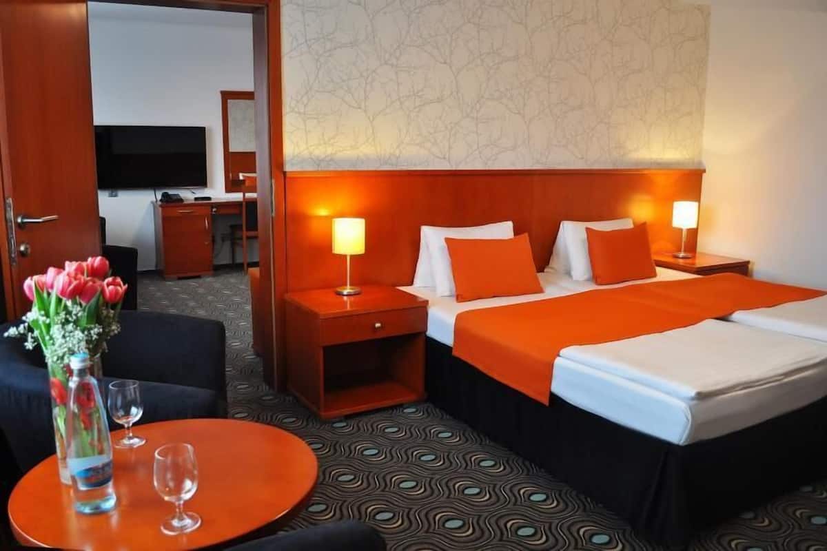 Une chambre d’hôtel avec un grand lit, deux tables de chevet avec des lampes, un bureau avec une chaise, une télévision et un vase avec des fleurs.