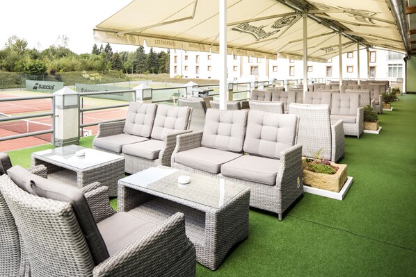 Une terrasse meublée avec des meubles en osier et, au fond, un court de tennis.
