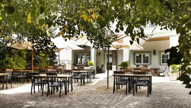 Un café al aire libre con mesas y sillas dispuestas en un patio empedrado, rodeado de árboles y un edificio con una decoración verde y blanca.