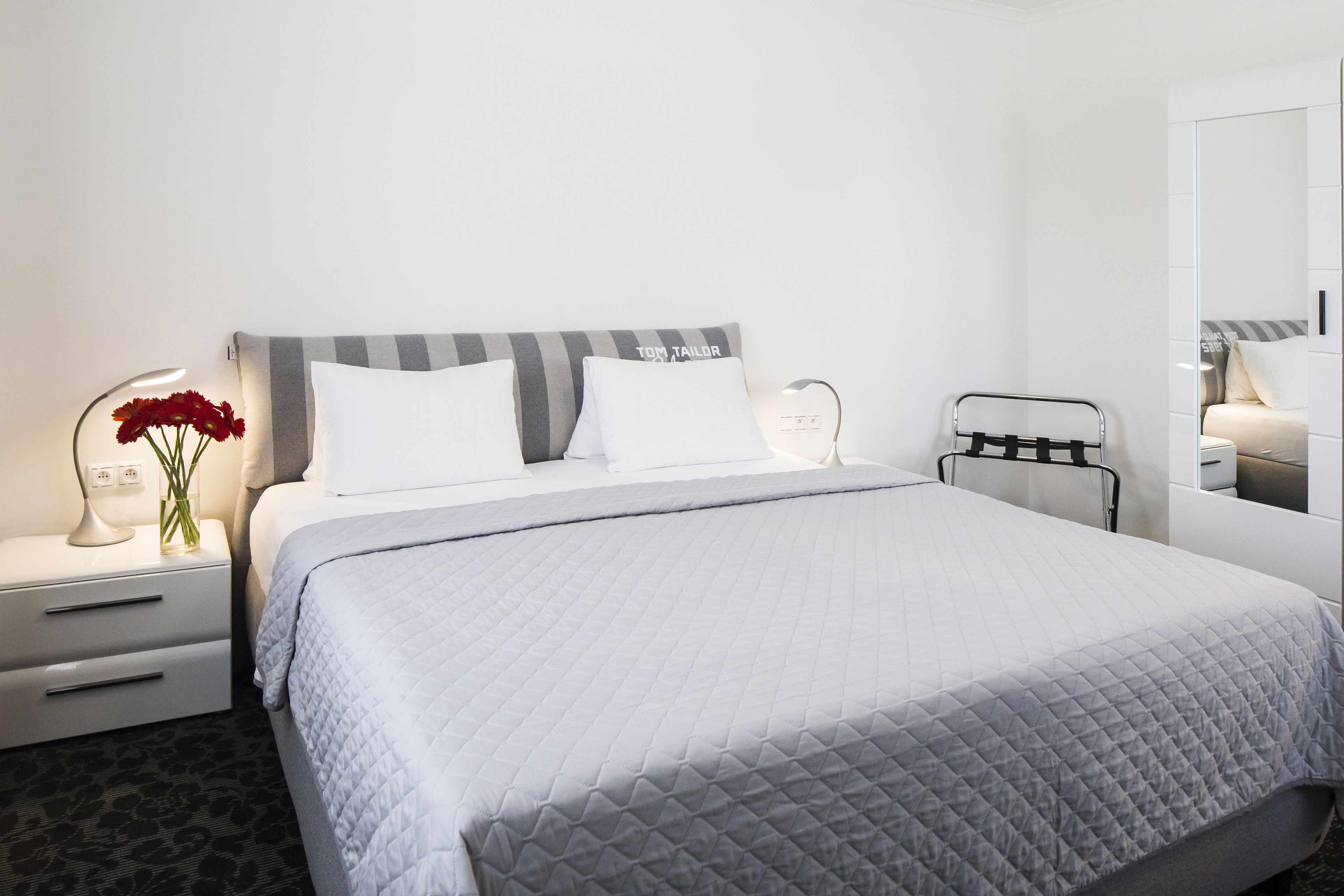 A neatly made bed with a grey quilt, a bedside table with a lamp and a vase of flowers, and a metal rack.