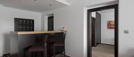 Habitación de hotel moderna con una barra de madera oscura, dos sillones acolchados, un pequeño estante con copas de vino, un cuadro en la pared y una alfombra a rayas.