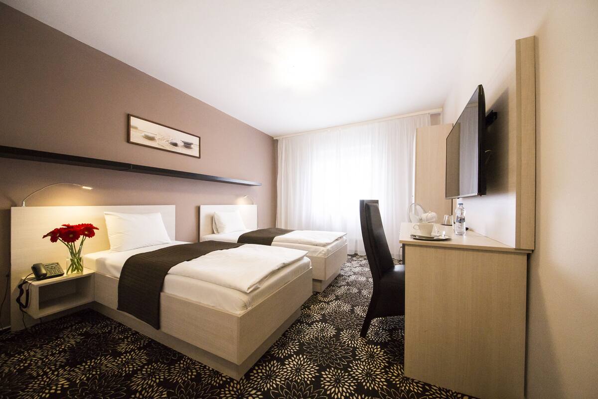 Une chambre d’hôtel avec deux lits, un bureau, une chaise, une télévision et un vase avec des fleurs.