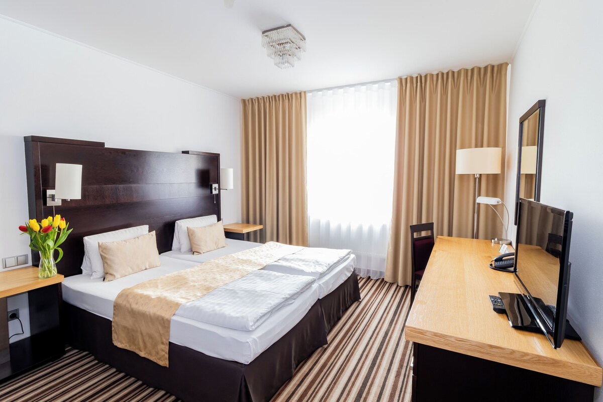 Ein Hotelzimmer mit einem großen Bett, einem Schreibtisch mit Computer, einem Stuhl, einer Lampe und einem Spiegel.
