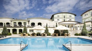 Una piscina con sillones y sombrillas, rodeada de un edificio con varios balcones y un cielo despejado.