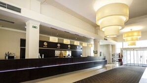 Zona de recepción de hotel con un mostrador oscuro, varios relojes y luminarias colgantes.