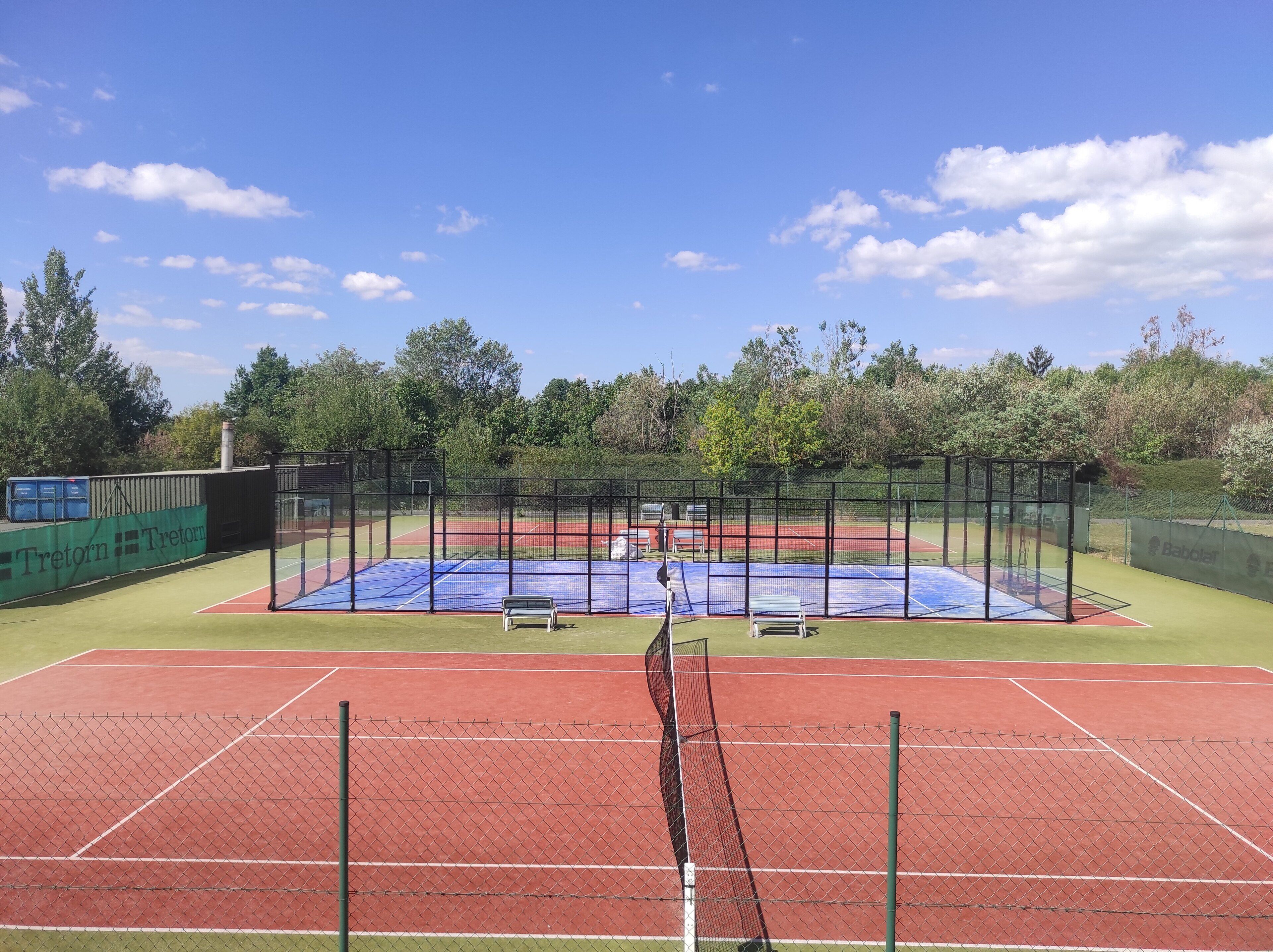 Een tennisbaan met net, omgeven door een hek en groen.