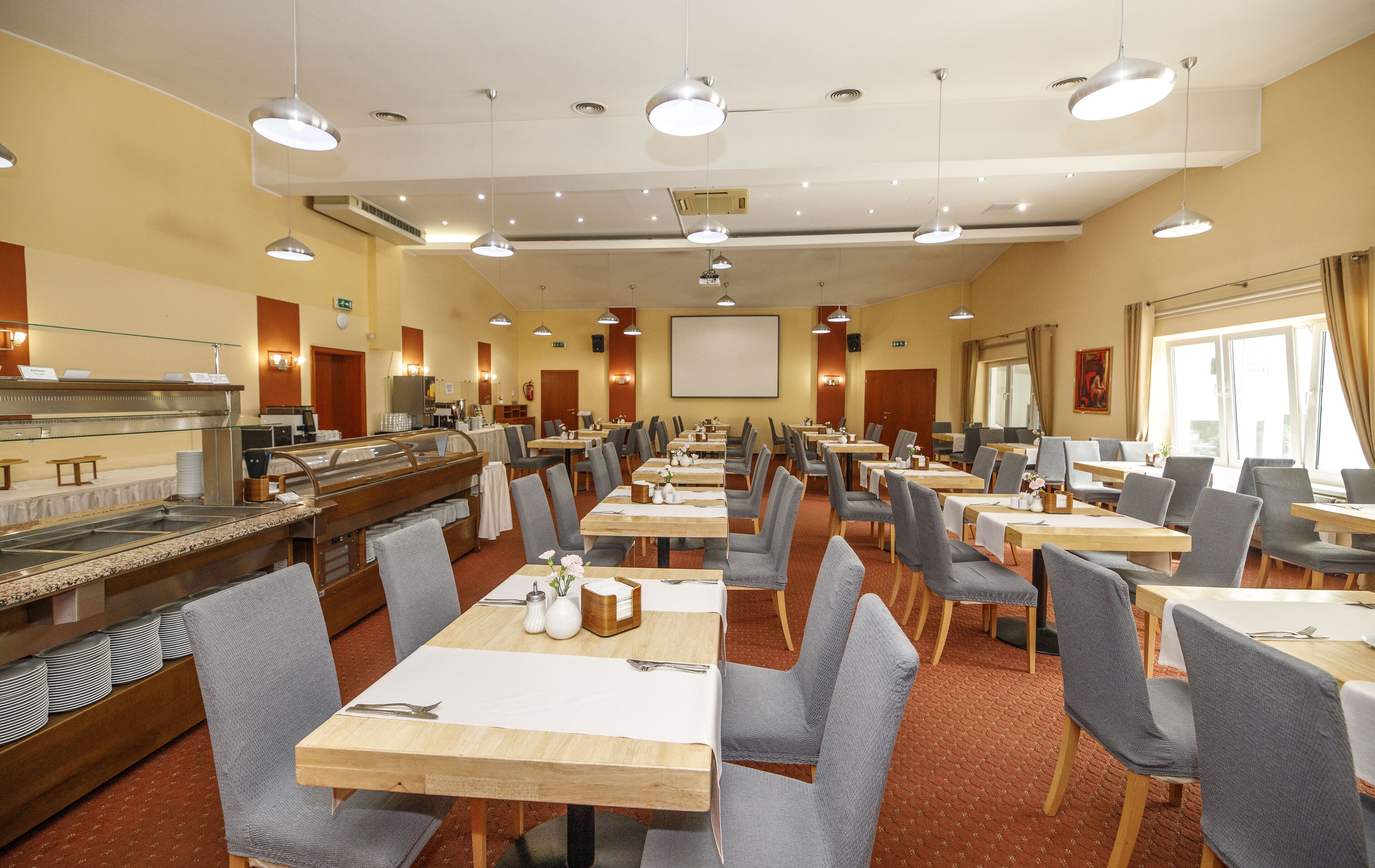 Une salle à manger d'hôtel avec de nombreuses tables et chaises, un buffet et un écran de projection fixé au mur.