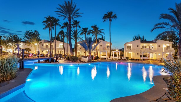 A swimming pool with a curved edge, surrounded by palm trees and a building with balconies.
