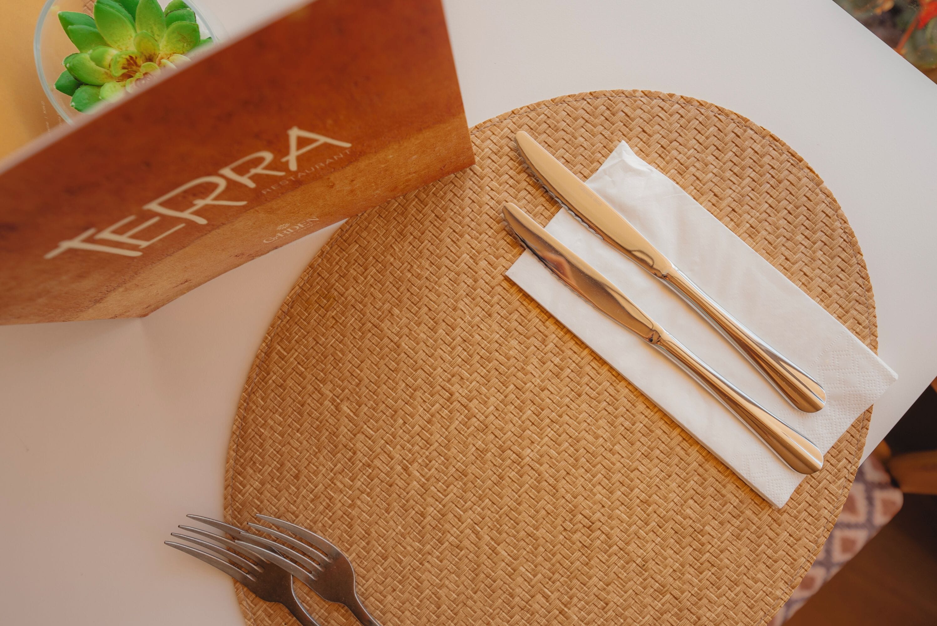 Un montaje para comer con un protector de mesa tejido, una servilleta blanca, un tenedor dorado y un cuchillo sobre una mesa blanca.