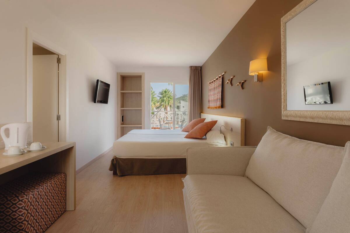 Une chambre d’hôtel avec un lit, un canapé, une télévision, un bureau avec une bouilloire et une vue sur des palmiers.