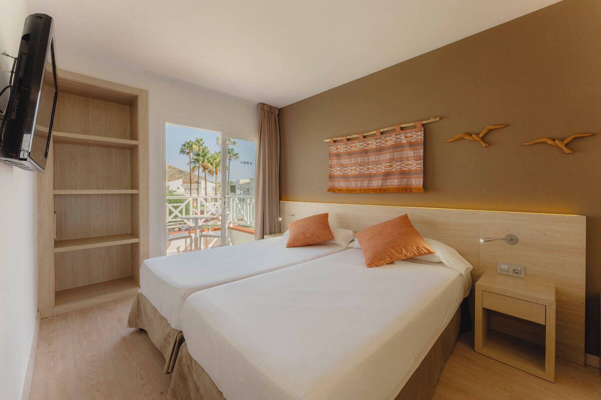 Une chambre d’hôtel avec deux lits, une armoire en bois, un téléviseur fixé au mur et un balcon offrant une vue sur les palmiers.