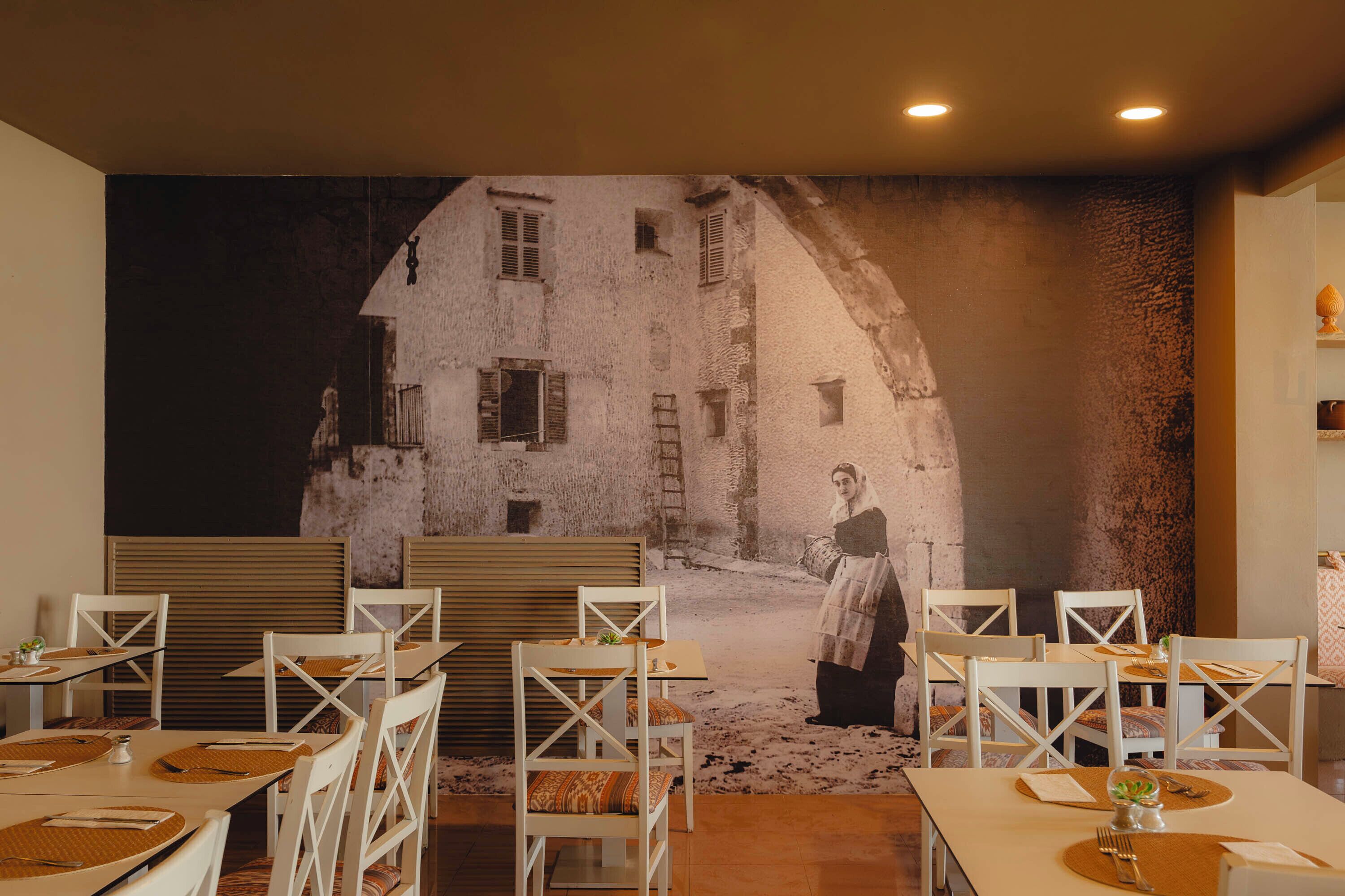 A dining area with white tables and chairs, a mural of a rustic building, and a woman standing in front of it.