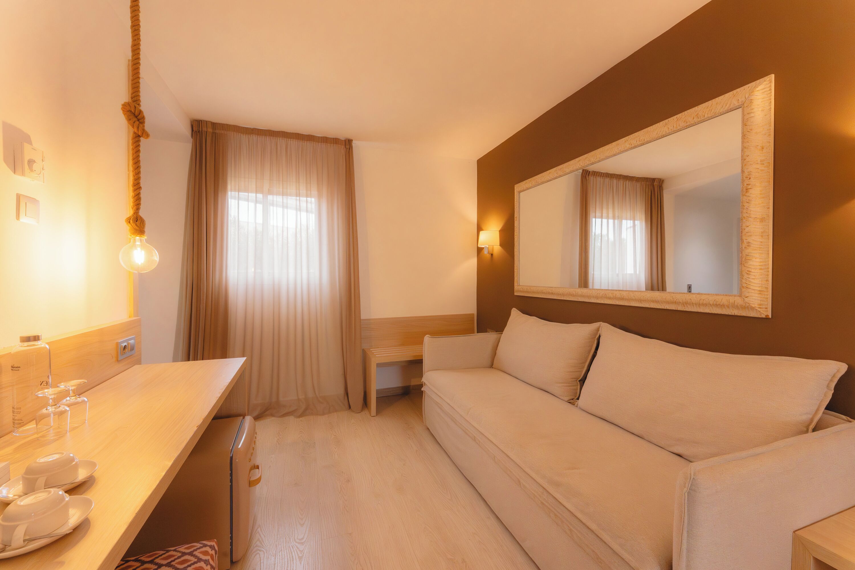 A hotel room with a wooden desk, a sofa, a mirror, and a hanging light fixture.