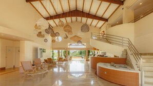 A spacious lobby with a high ceiling, wooden beams, and hanging wicker lamps. There is a reception desk and a staircase to the upper level.