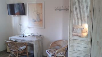 Ein Zimmer mit weißem Schreibtisch, zwei Korbstühlen, einem an der Wand befestigten Fernseher und einem Spiegel mit Holzrahmen.