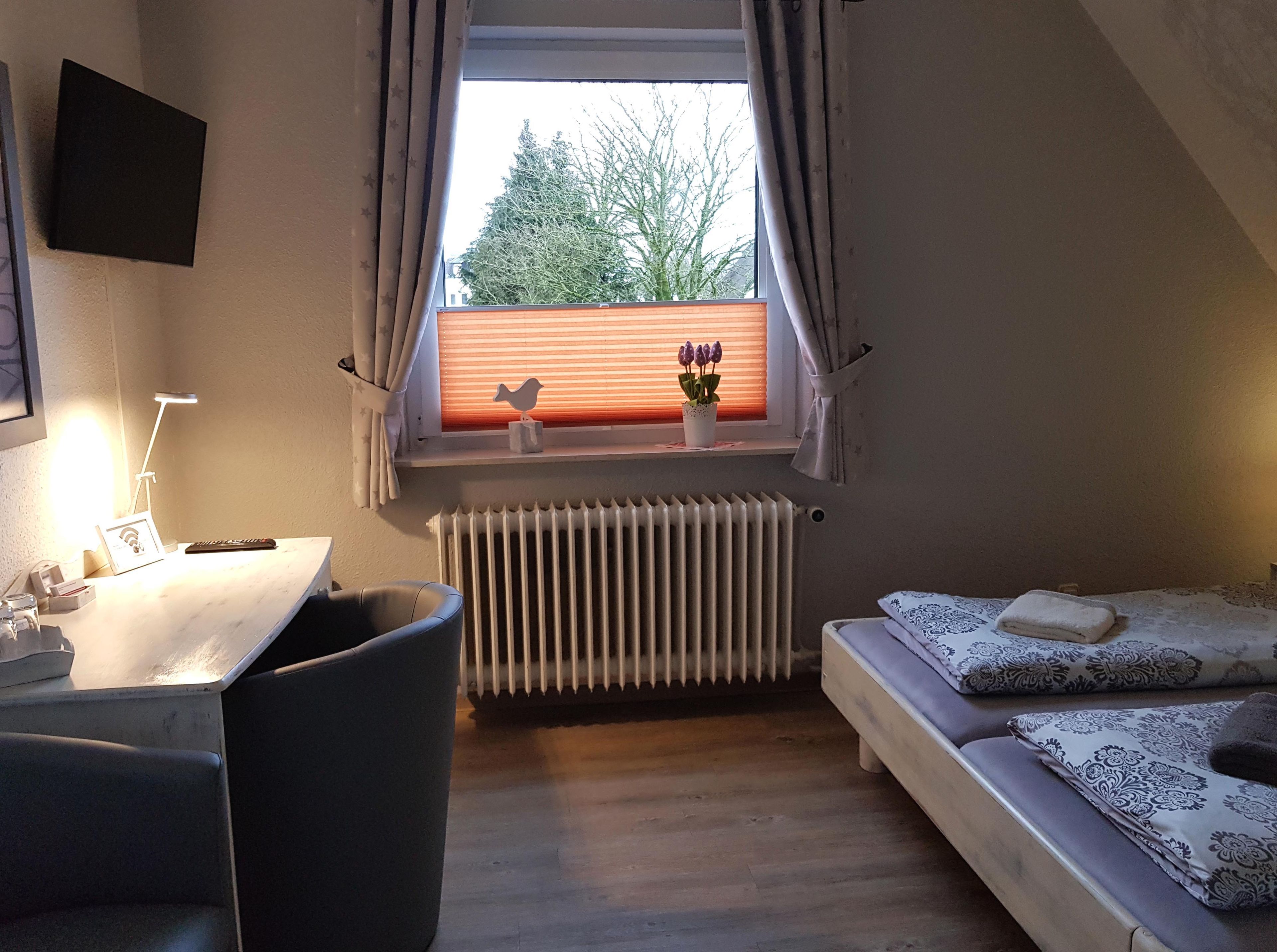 A small, cozy room with a desk, a chair, a radiator, a window with curtains, and a bed with patterned bedding.