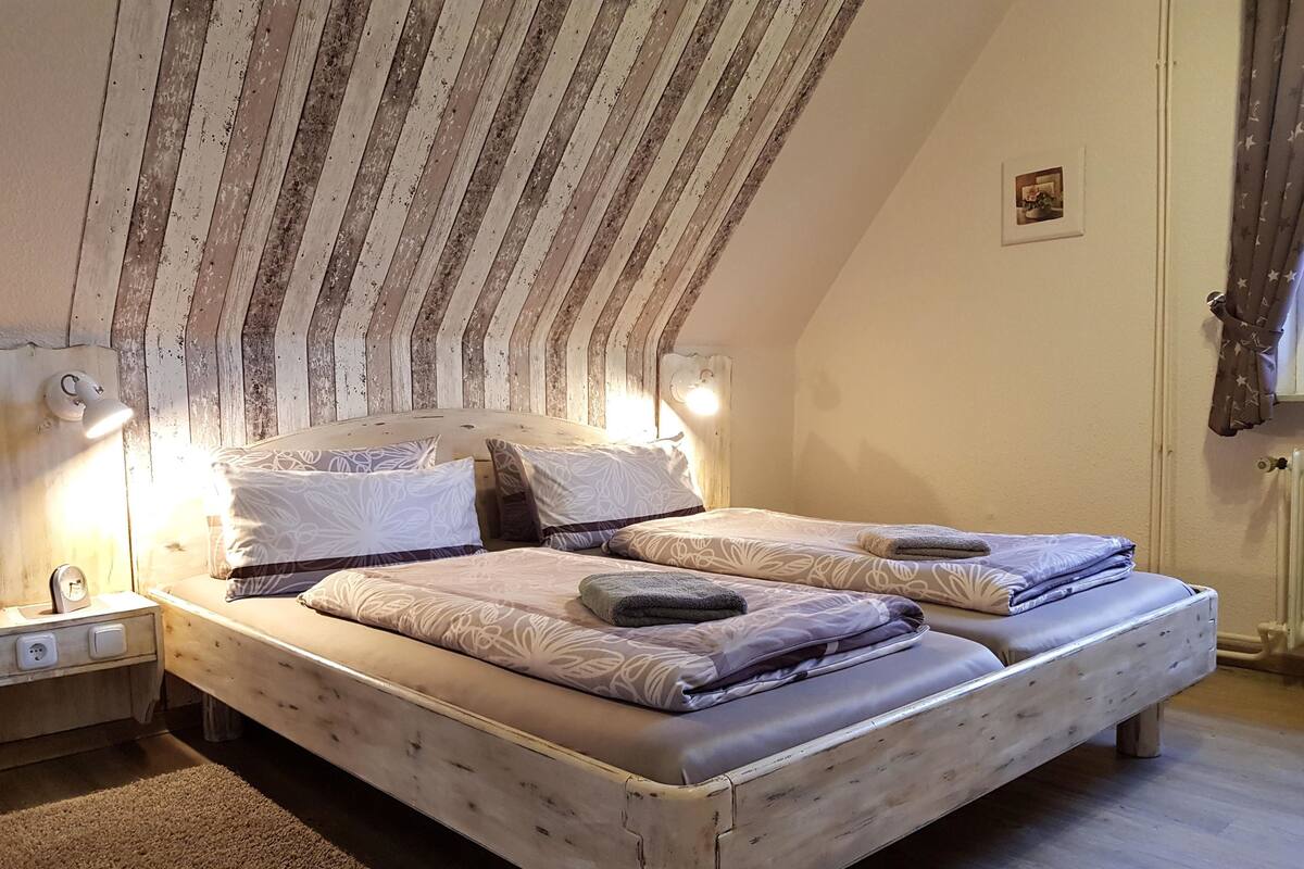 Une chambre avec un plafond en pente, un cadre de lit en bois, deux tables de chevet avec des lampes et un petit tableau accroché au mur.