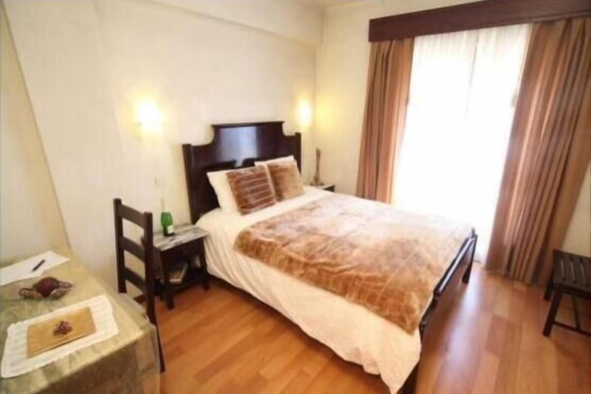 Une chambre à coucher avec un grand lit, une tête de lit en bois, deux tables de chevet, une commode, un bureau avec une chaise et une fenêtre avec des rideaux.