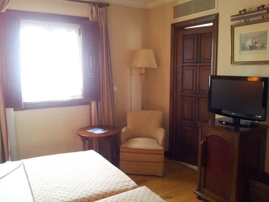 A hotel room with a bed, a chair, a TV, a lamp, and a window with curtains.
