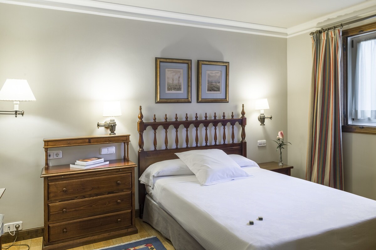 A bedroom with a wooden bed, two bedside tables, a nightstand, a lamp, a framed picture, and a window with curtains.