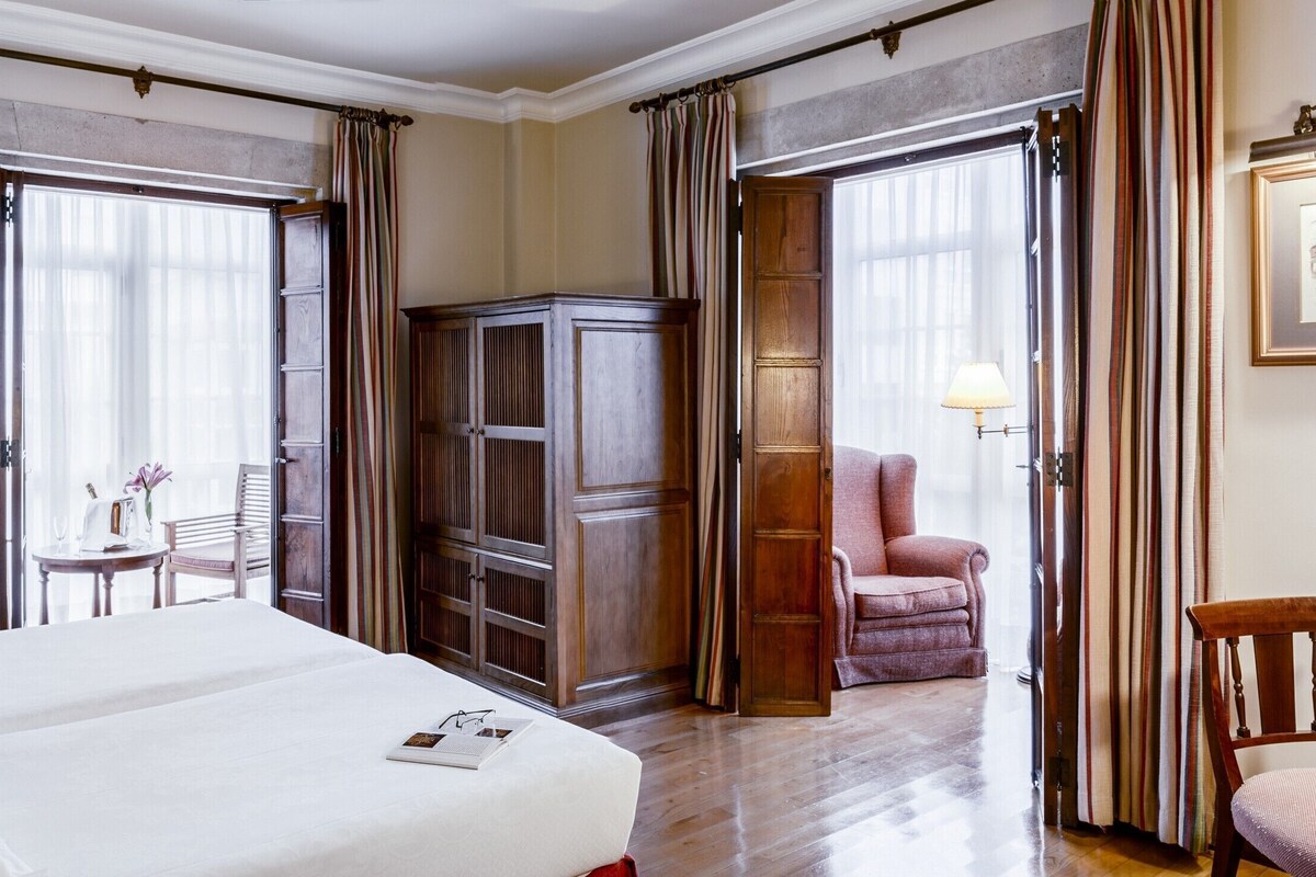 A hotel room with a large bed, a wooden wardrobe, a desk with a chair, and a window with curtains.