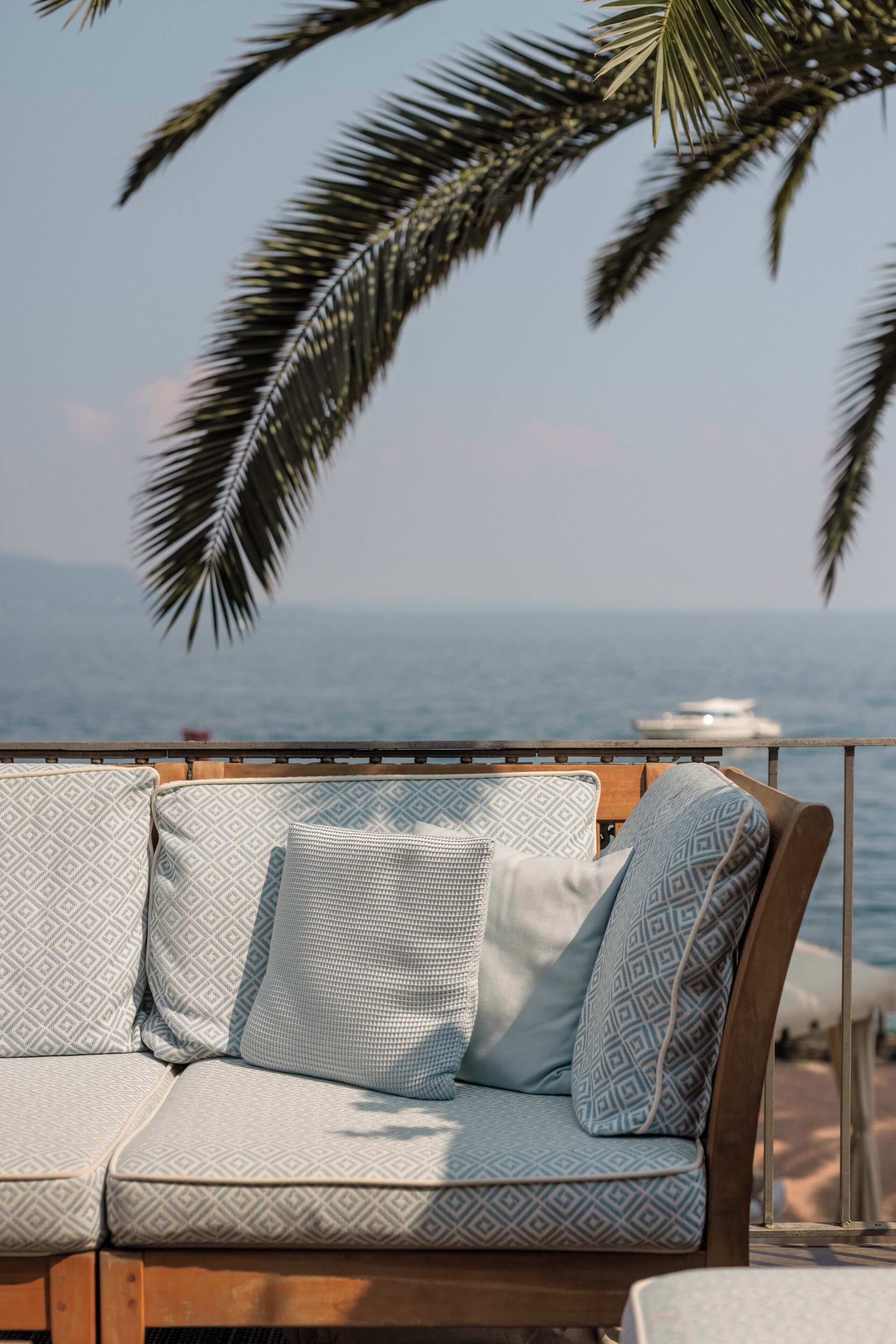 A wooden outdoor seating area with patterned cushions, overlooking a body of water and palm trees.