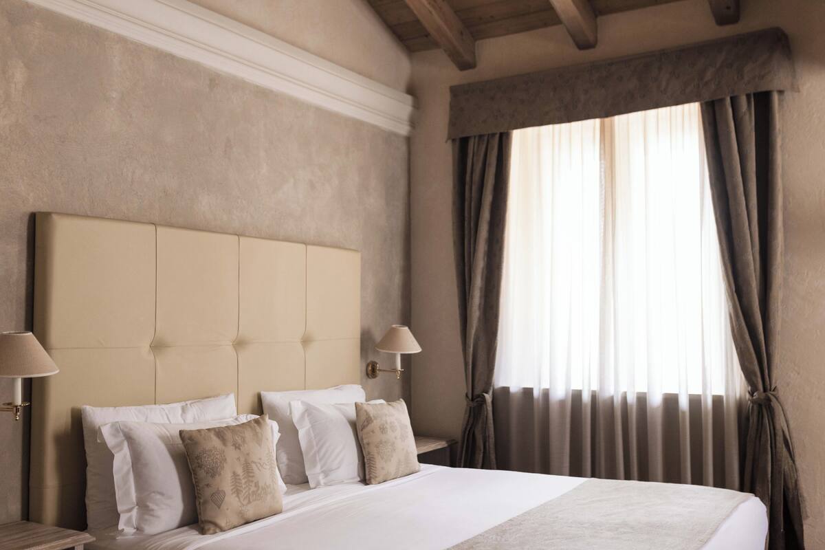 Une chambre d’hôtel avec un grand lit, deux tables de chevet, une lampe fixée au mur et une fenêtre avec des rideaux.