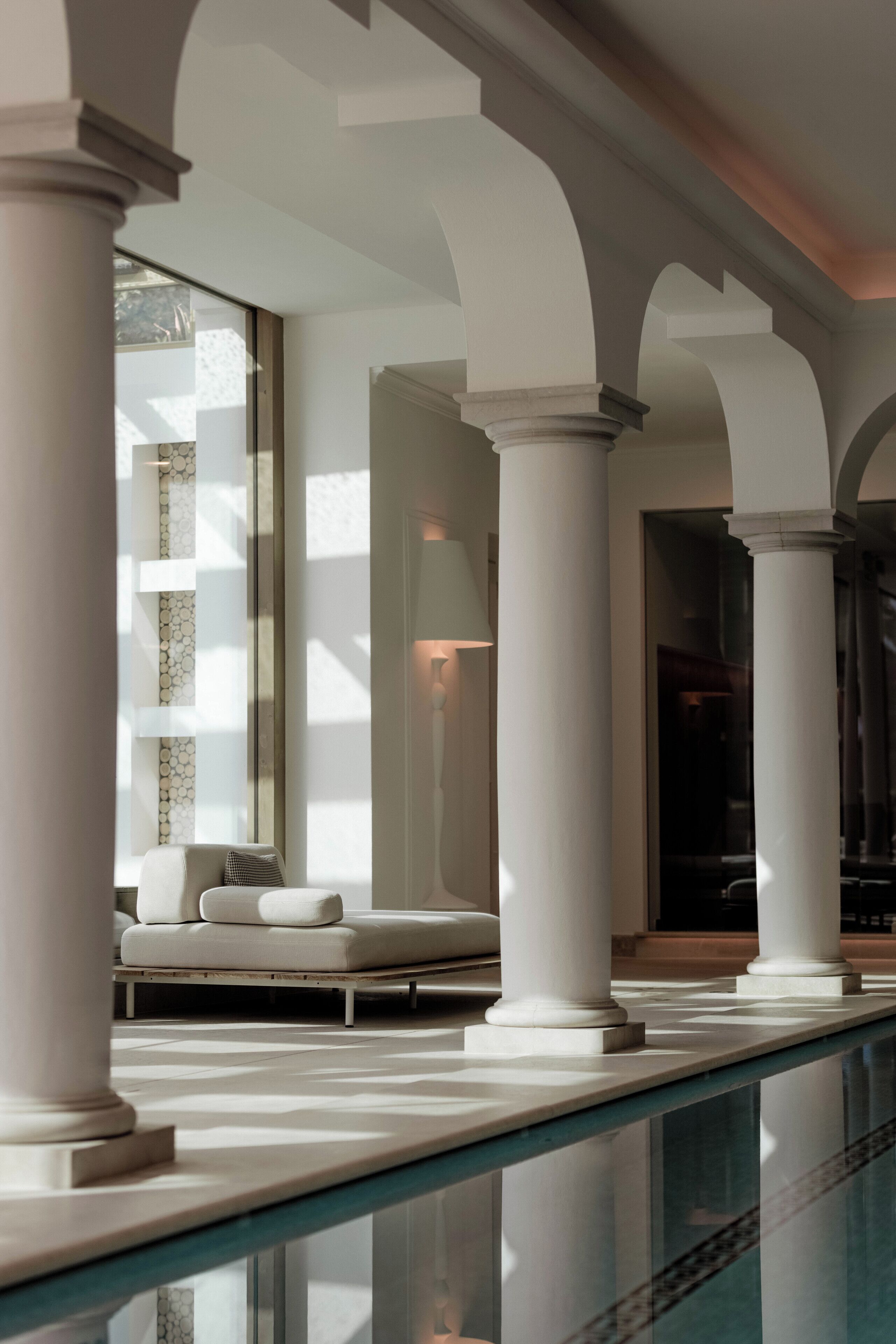 Un área de piscina interior moderna con columnas blancas, barandilla de cristal y un espacio de descanso con sofá y almohadas.