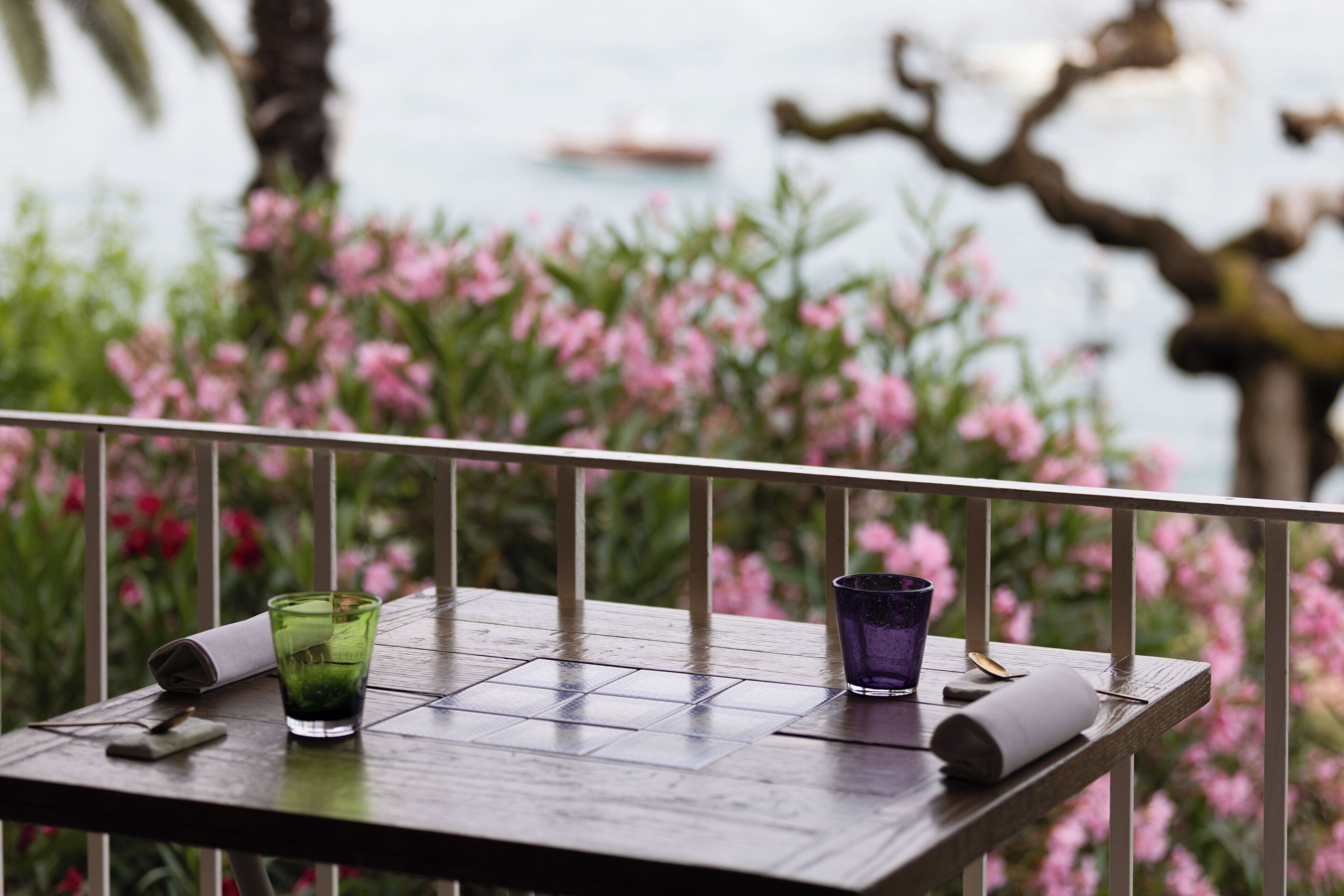 Ein Holztisch mit zwei Gläsern und einem Servietttuch, vor einer Kulisse aus rosafarbenen Blumen und einem Gewässer.