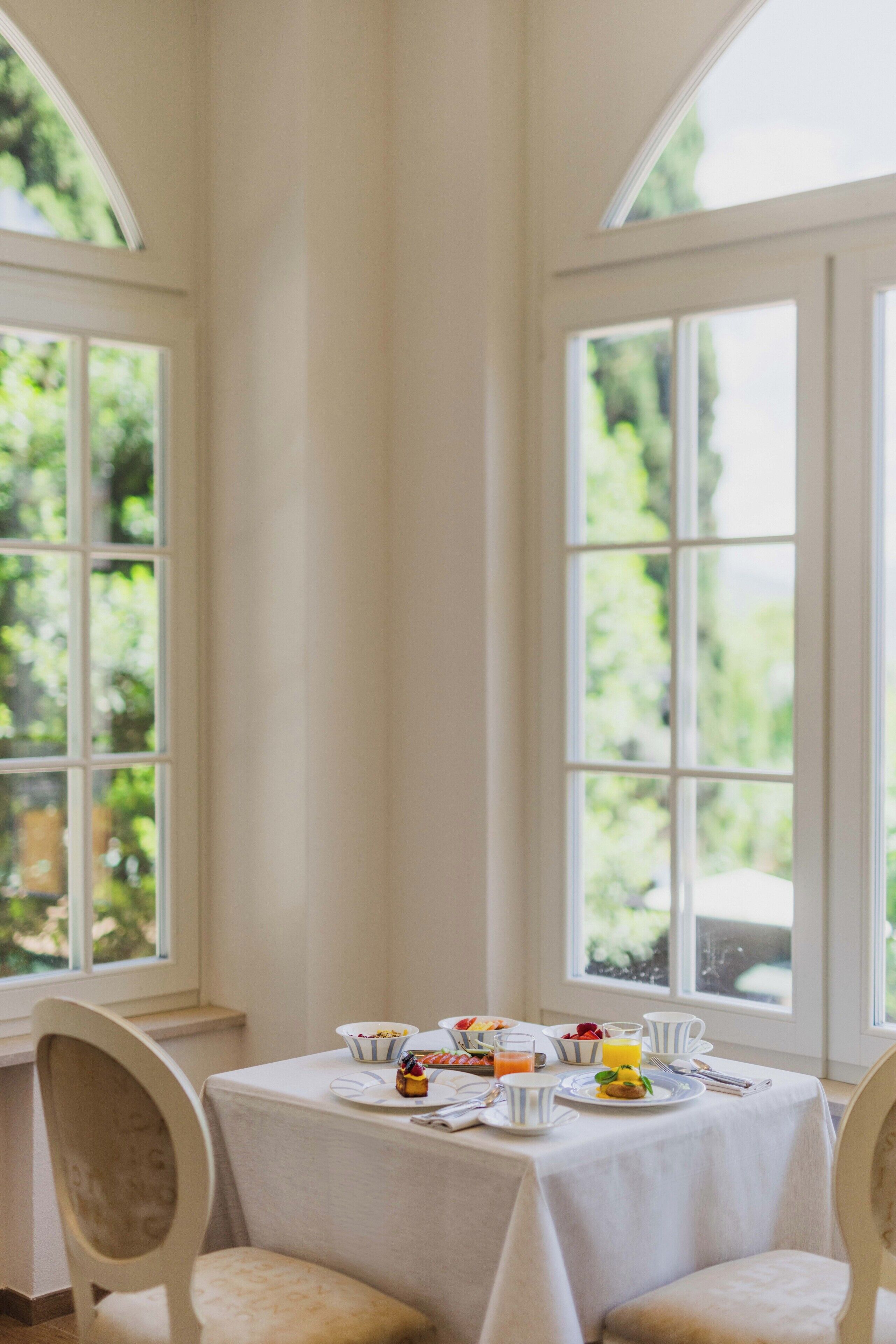 A breakfast table with various food items and drinks, set against a backdrop of large windows with a view of greenery.