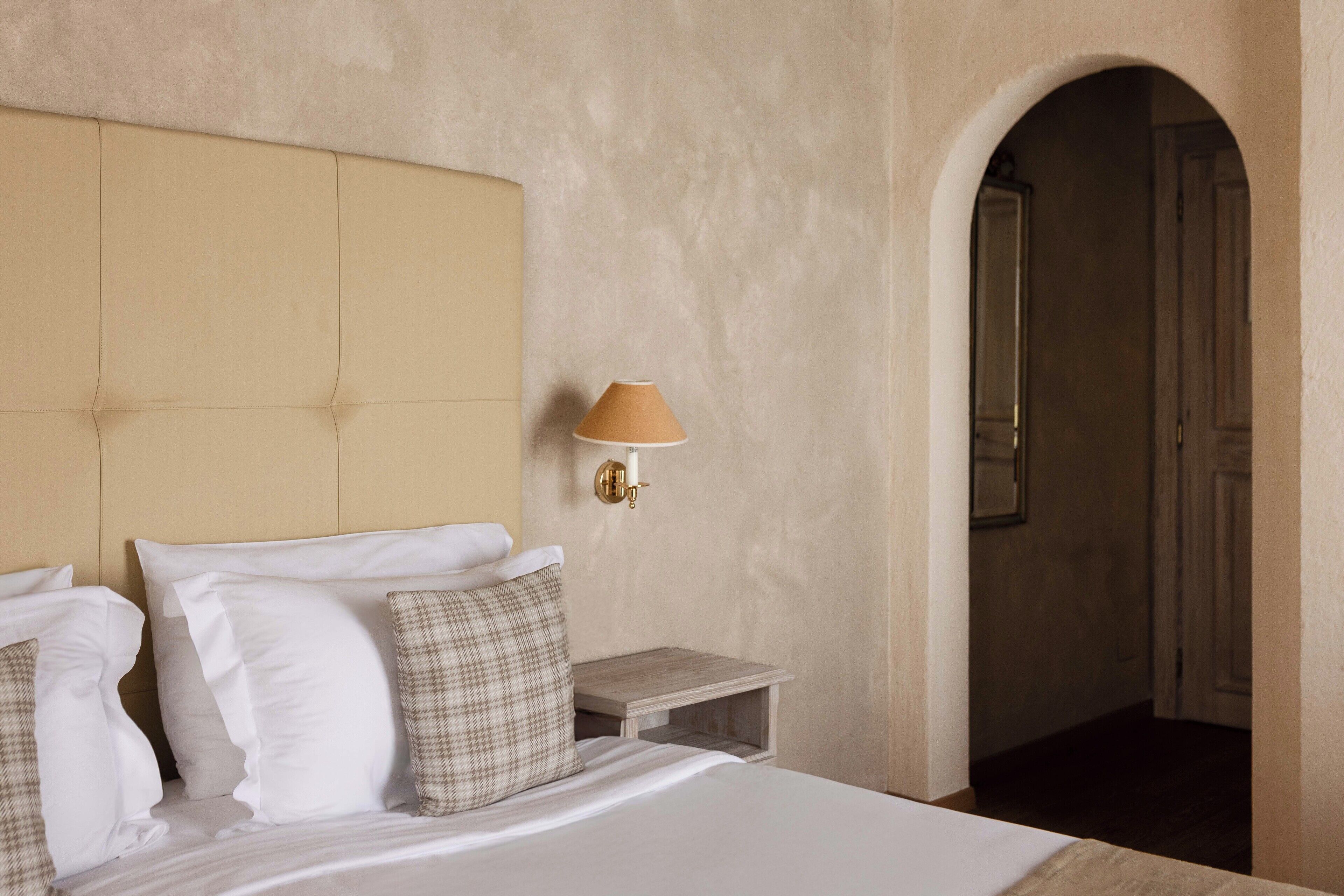 A hotel room with a large bed, a headboard, a nightstand with a lamp, and an arched doorway leading to another room.