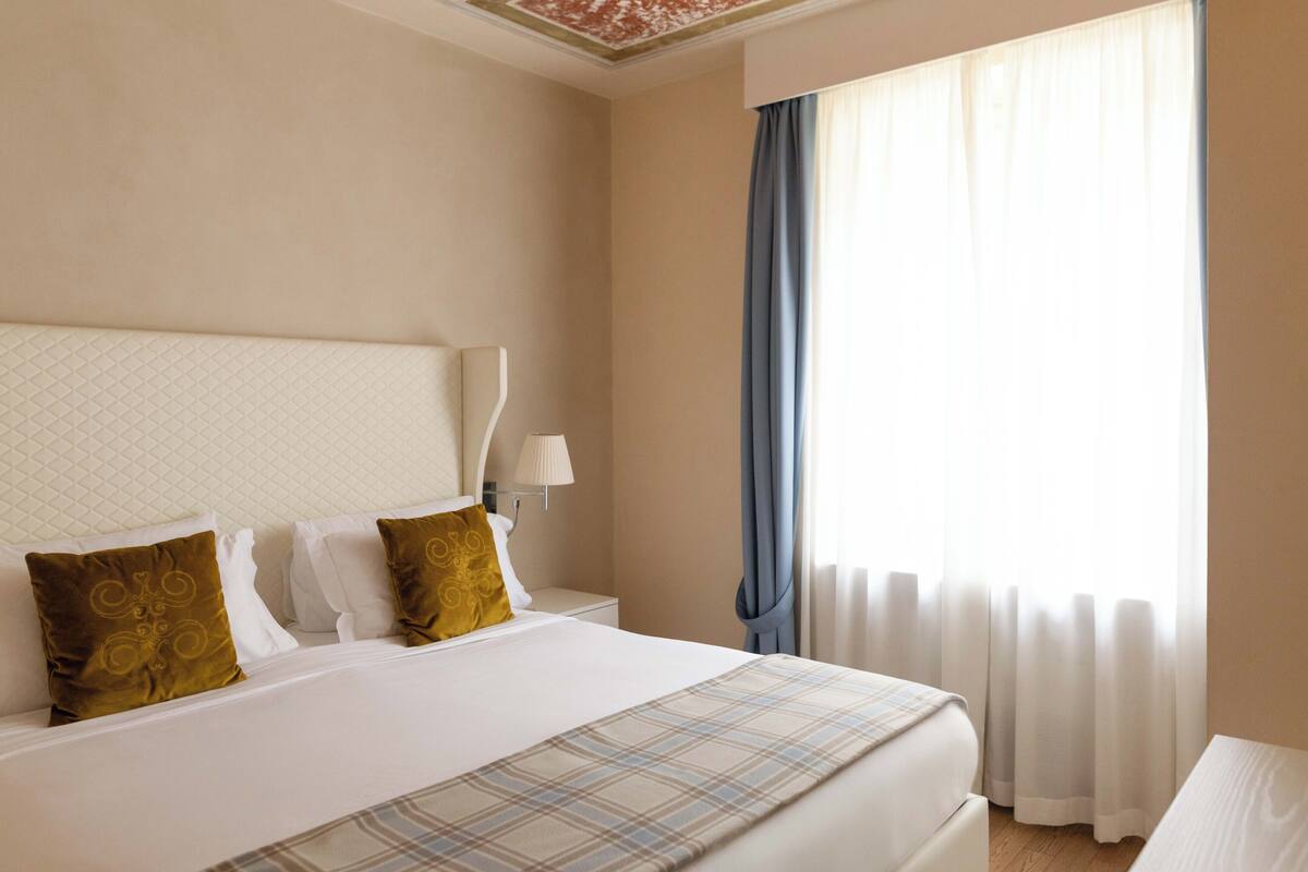 Une chambre d’hôtel avec un grand lit, une table de chevet, une lampe et une fenêtre avec des rideaux.