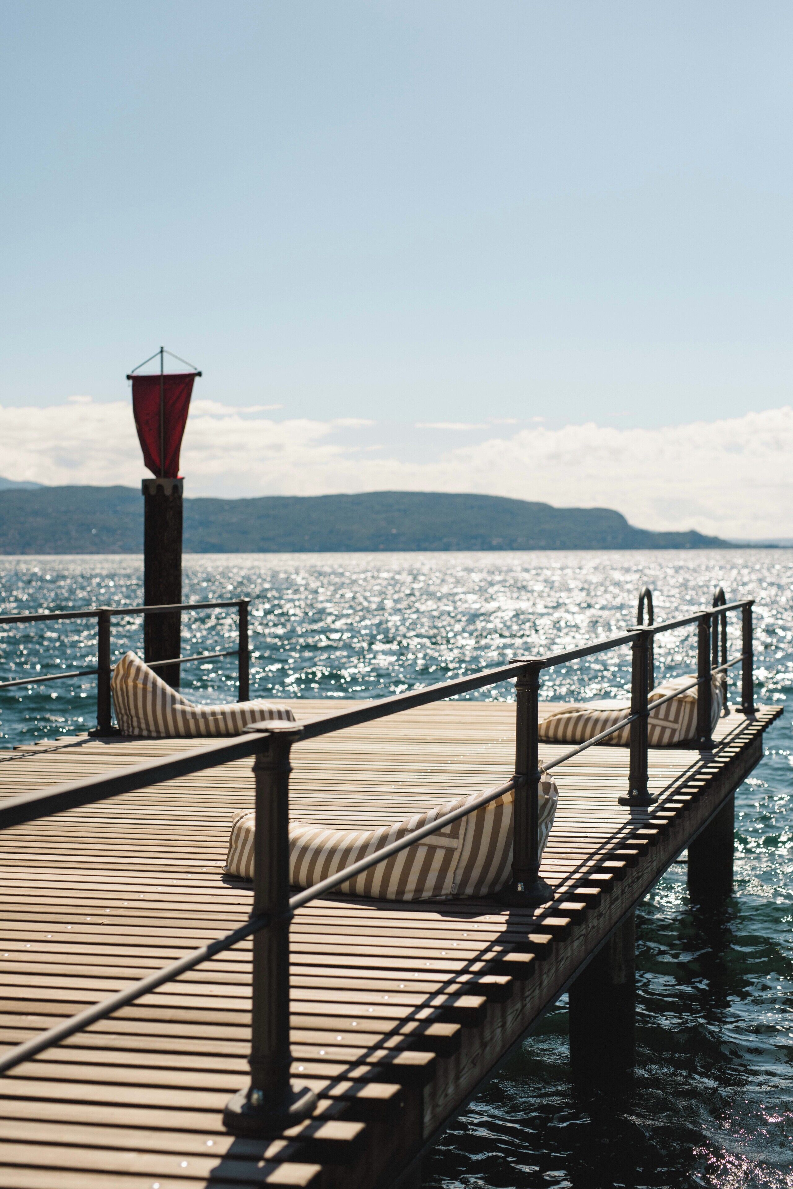 Un muelle de madera con cojines a rayas y un farolillo rojo, que conduce a una masa de agua con una cadena montañosa distante.