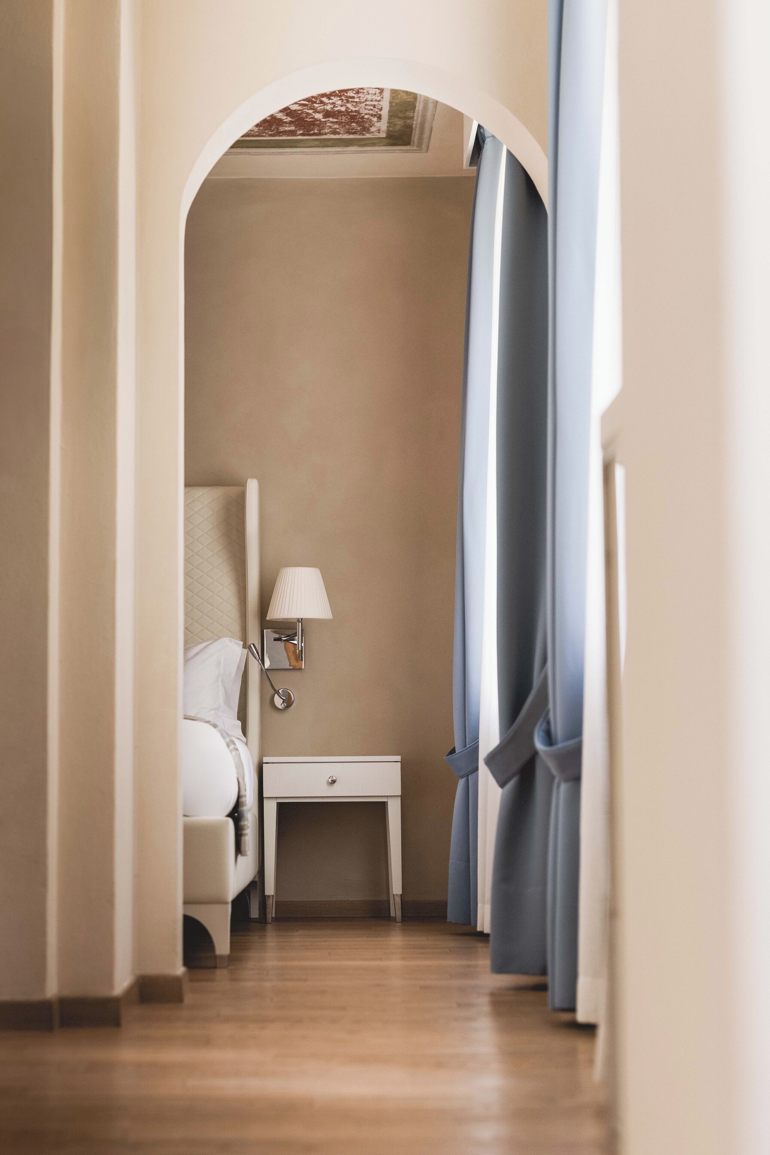 A narrow hallway with a bed, a nightstand, a lamp, and a window with curtains.