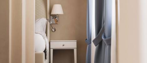 A narrow hallway with a bed, a nightstand, a lamp, and a window with curtains.