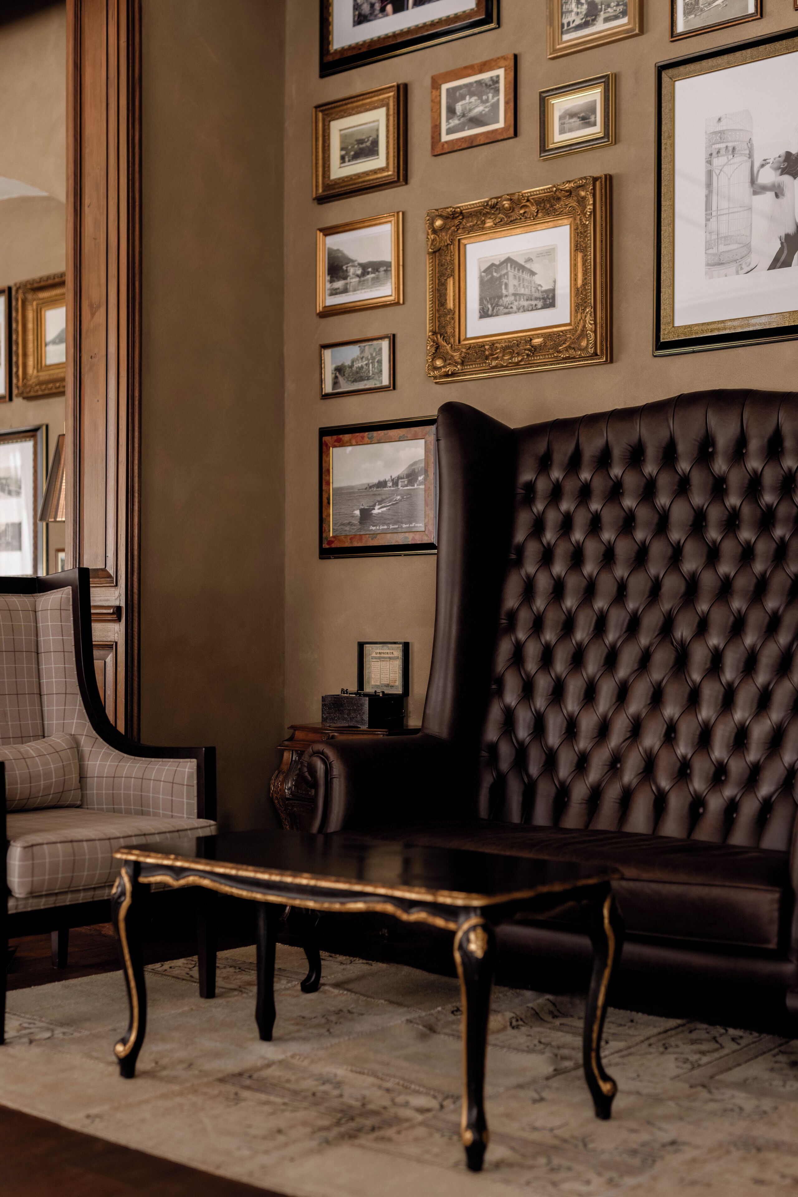 A room with a dark brown tufted sofa, a matching armchair, and a small table. The walls are adorned with framed pictures.