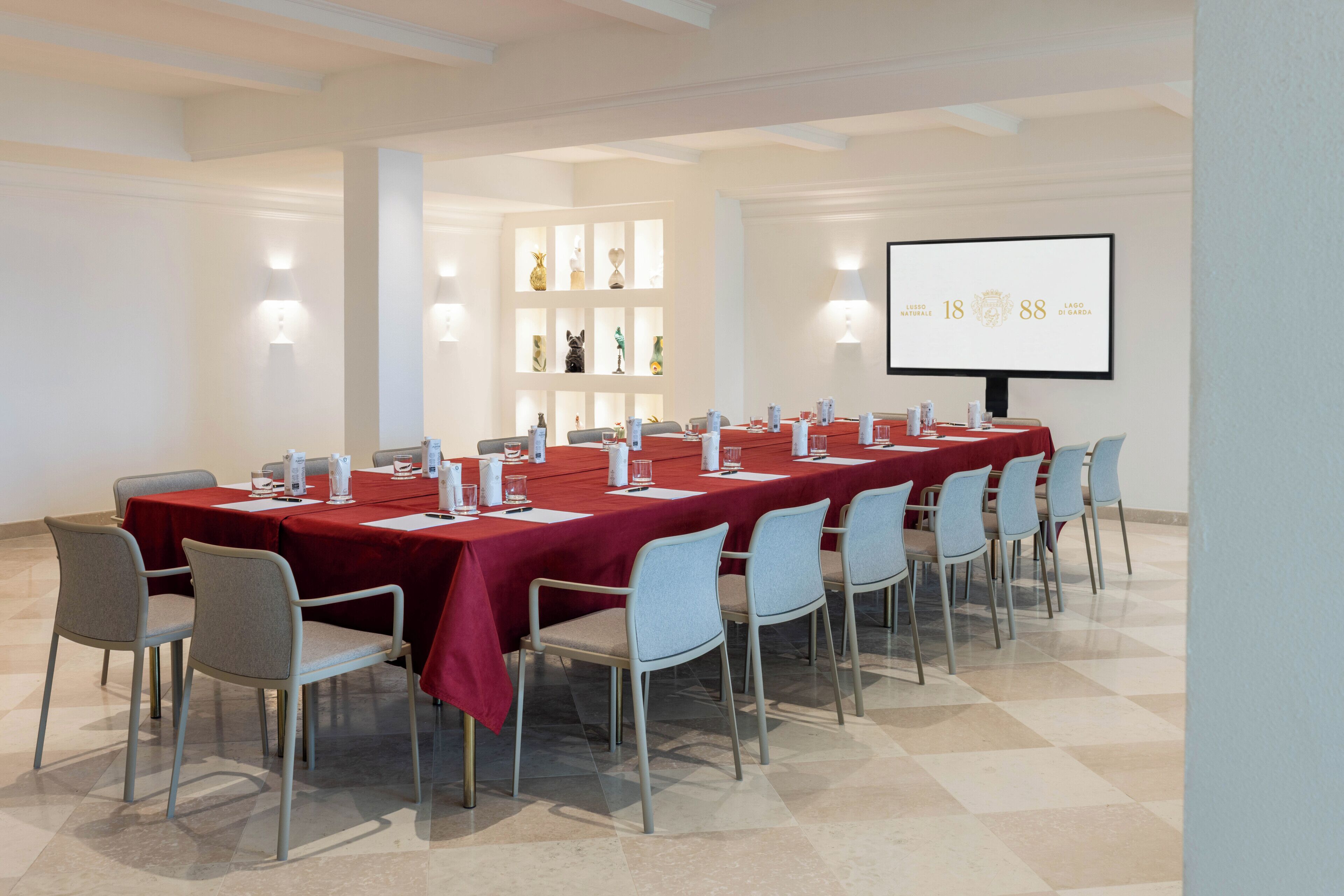 Un salón de conferencias con una larga mesa, sillas, una pantalla de proyector y estanterías con objetos decorativos.