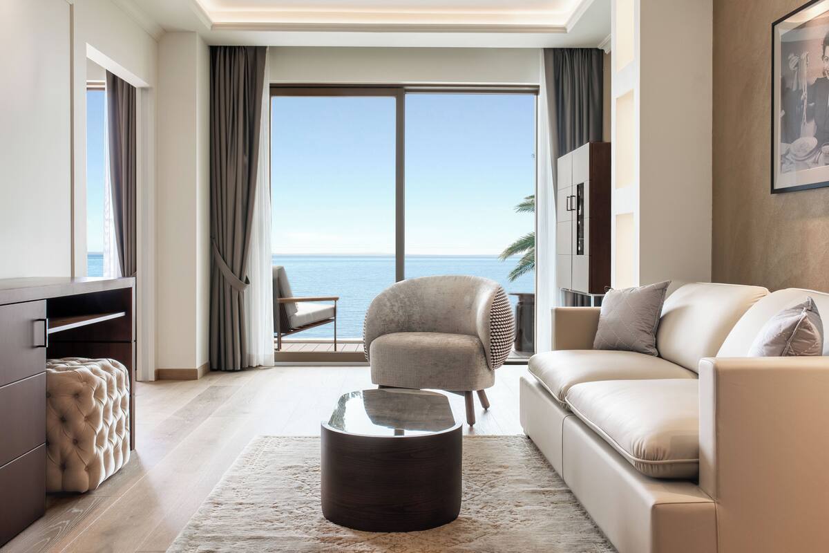 Un salon moderne comprenant un canapé, un fauteuil et un pouf, un téléviseur à écran plat et une grande fenêtre donnant sur la mer.