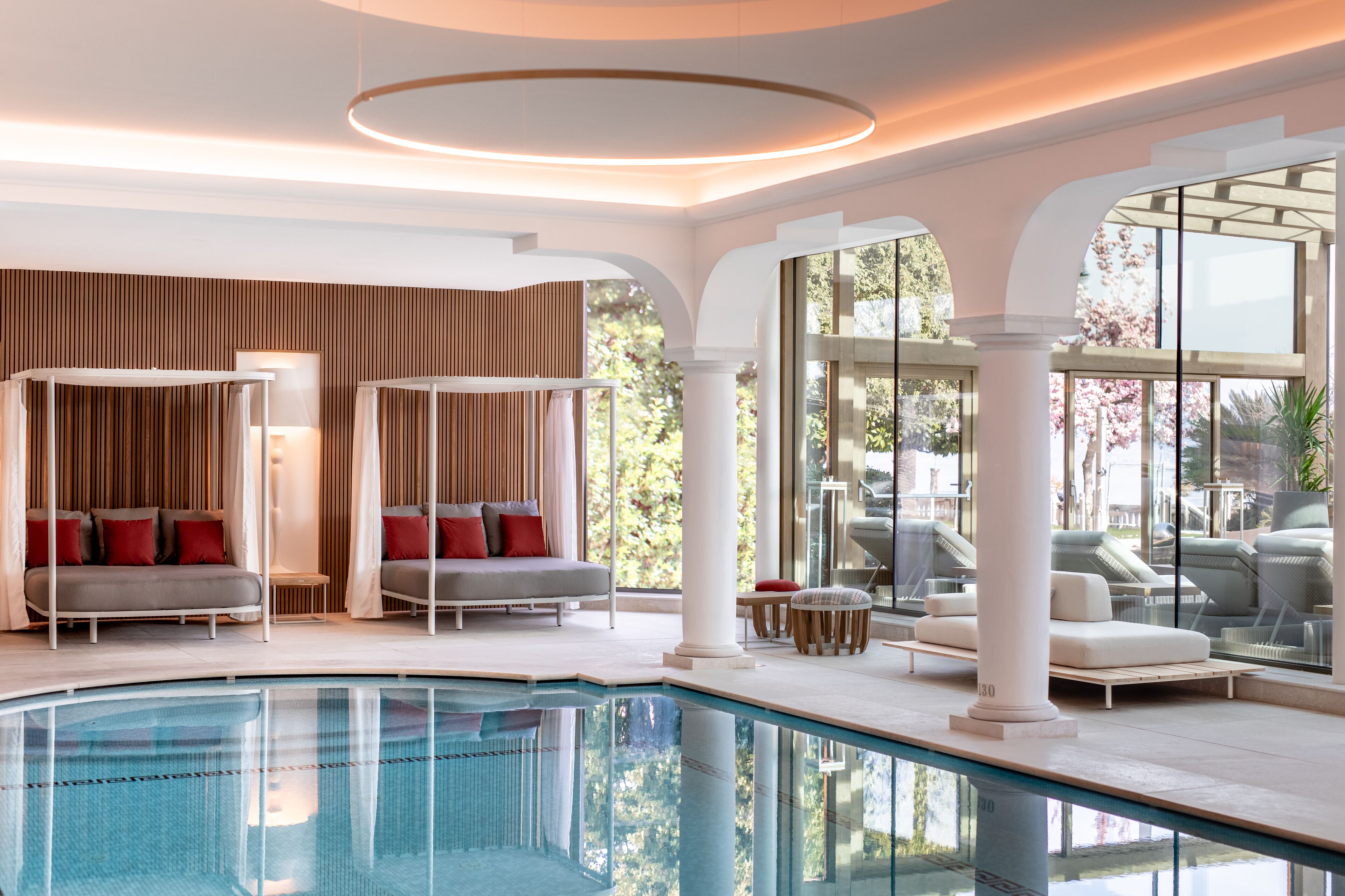A modern indoor pool area with a glass enclosure, white columns, and seating arrangements.