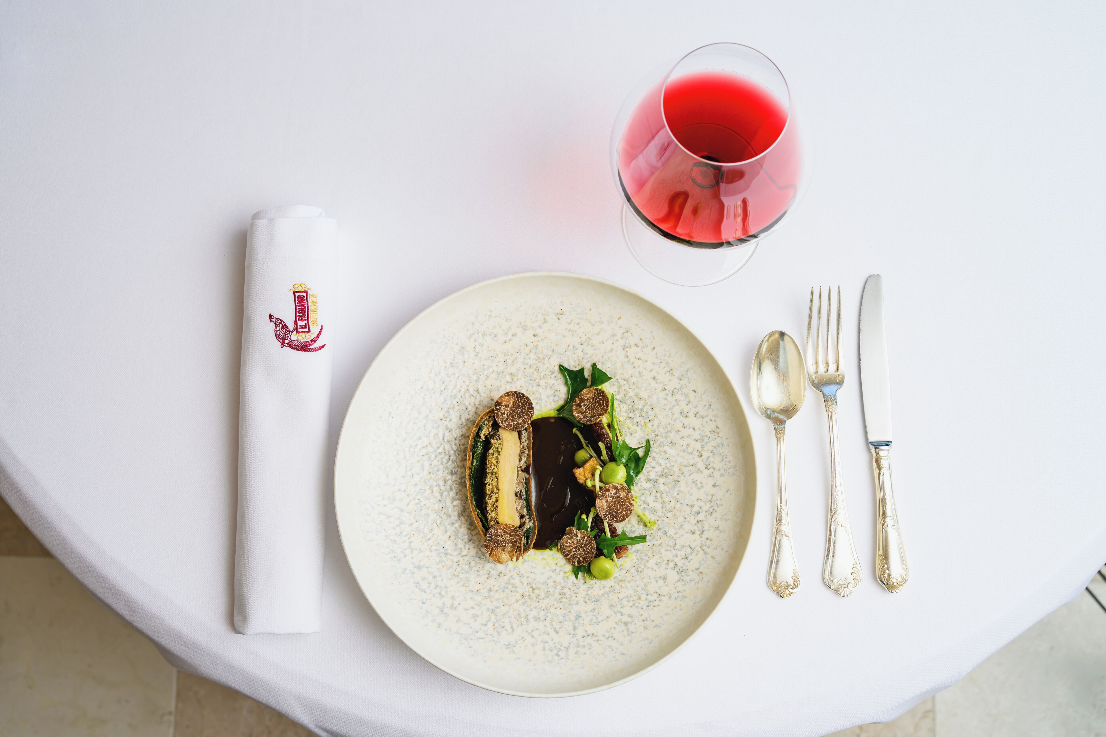 A white table with a plate of food, a glass of red wine, and silver cutlery.