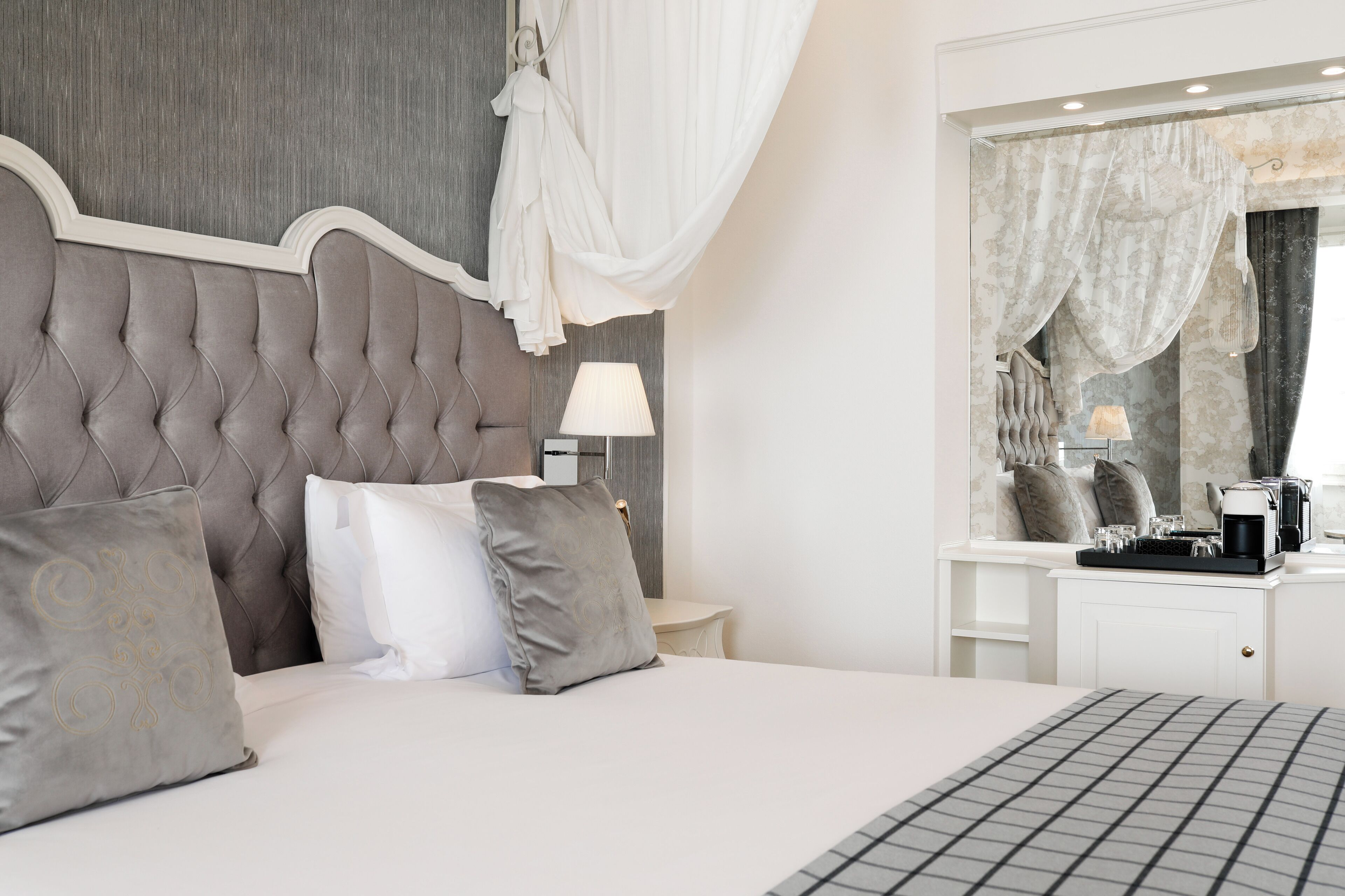Une chambre à coucher avec une tête de lit capitonnée, un lit avec une literie blanche et grise, une table de chevet avec une lampe et une petite table de nuit avec une machine à café.